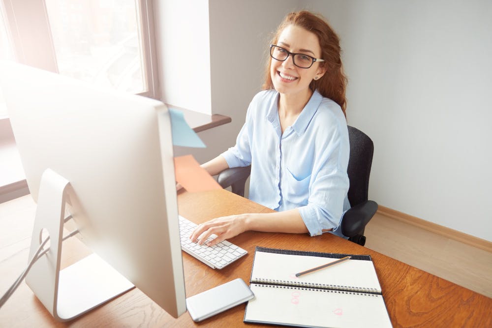 Eine Frau mit Brille sitzt an einem Schreibtisch vor einem Computer und lächelt in die Kamera.