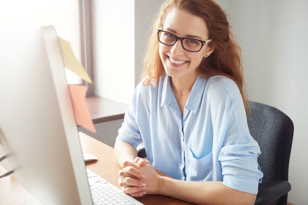 Eine lächelnde Frau sitzt vor einem Schreibtisch mit einem Monitor, trägt eine Brille und hat ihre Hände auf dem Schreibtisch.