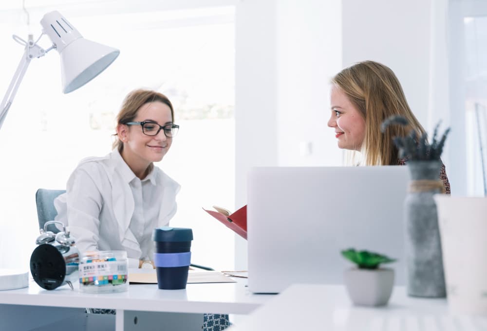 Zwei Frauen unterhalten sich an einem Schreibtisch in einem sauberen Büro.