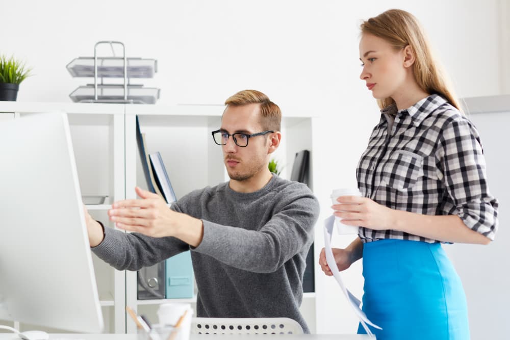 Ein Mann mit Brille zeigt einer Frau, die hinter ihm steht, etwas auf seinem Monitor