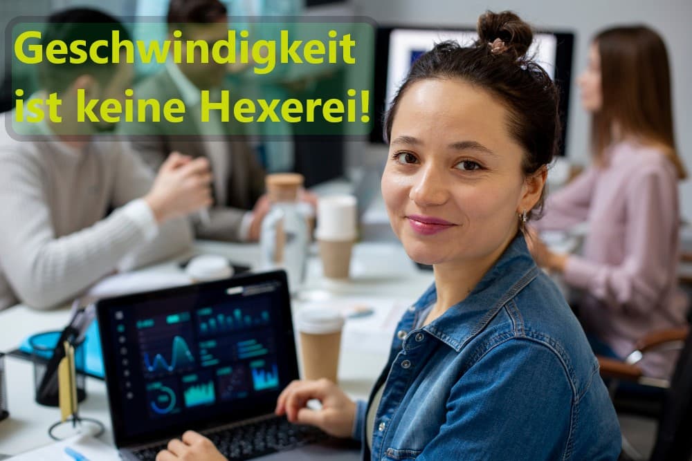 Eine Frau sitzt mit einer Gruppe an einem Schreibtisch mit einem Laptop und schaut in die Kamera.