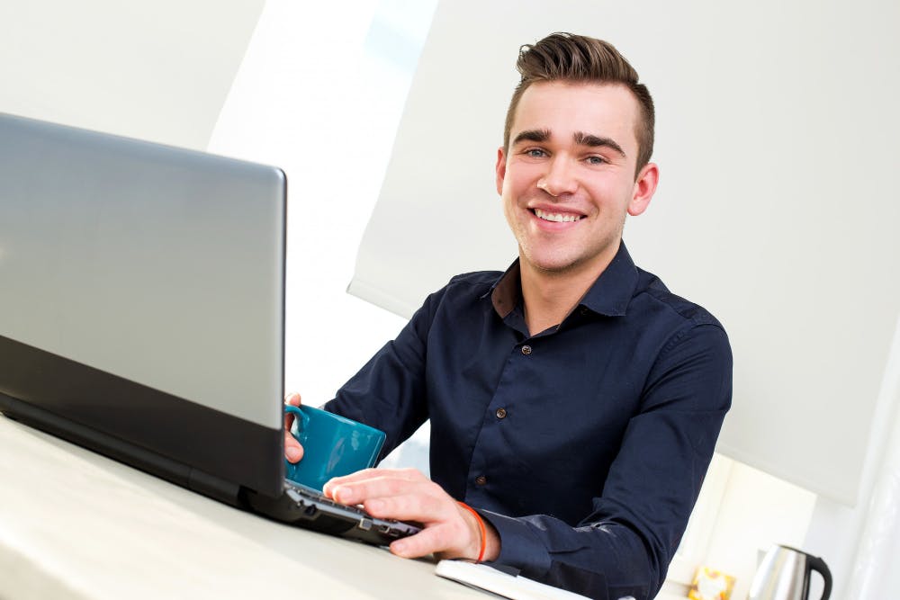 Ein Mann sitzt mit einer Tasse vor seinem Laptop und lächelt in die Kamera.