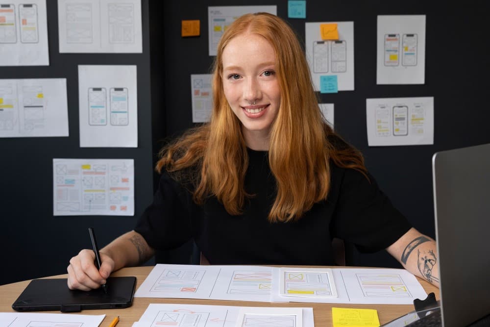 Eine Frau sitzt an ihrem Schreibtisch mit einem Grafik Tablet und einem Laptop, umringt von Grafiken.