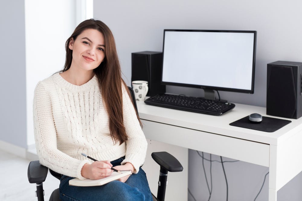 Eine lächelnde Frau sitzt vor einem Schreibtisch mit einem Computer, auf einem Schreibtischstuhl, hält einen Notizblock in der Hand und schaut in die Kamera.
