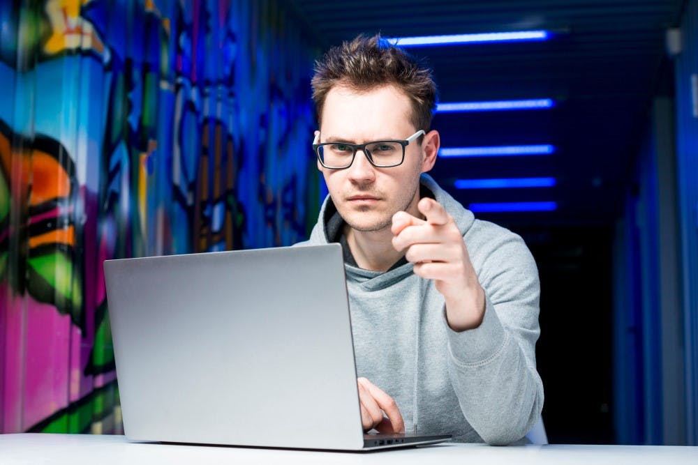 Ein Mann sitzt vor einem Laptop während er direkt in die Kamera schaut und mit dem Finger zeigt.