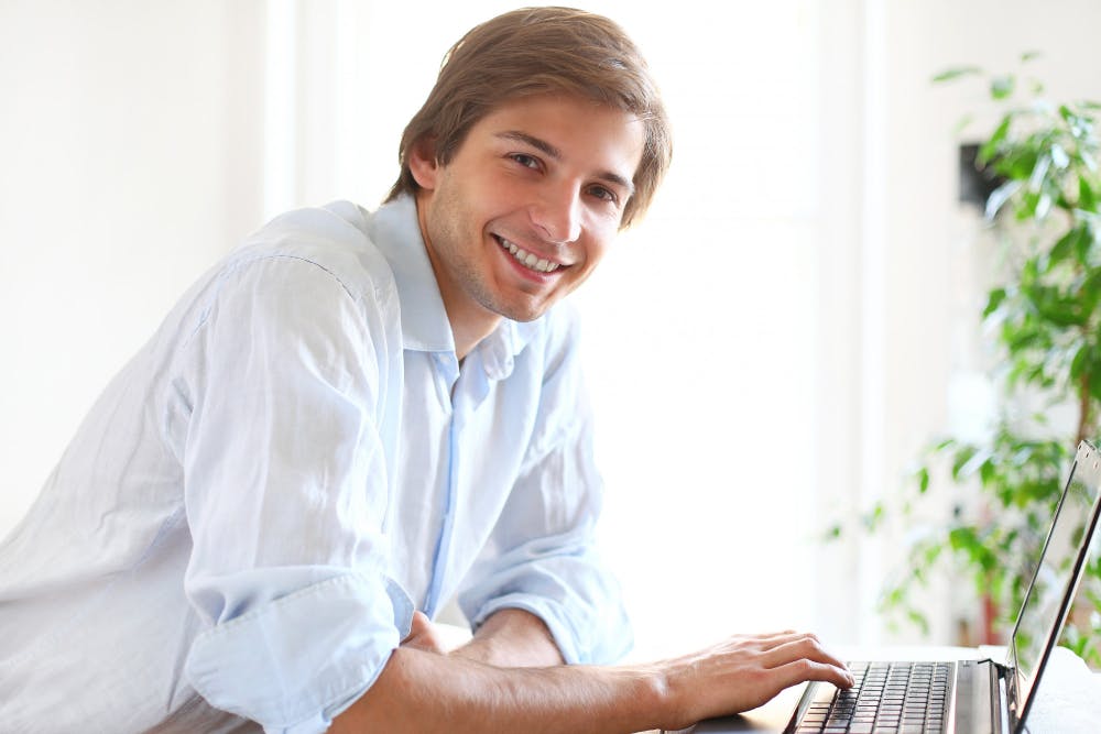Ein Mann sitzt nach vorne gelehnt an seinem Laptop und schaut lächelnd in die Kamera.