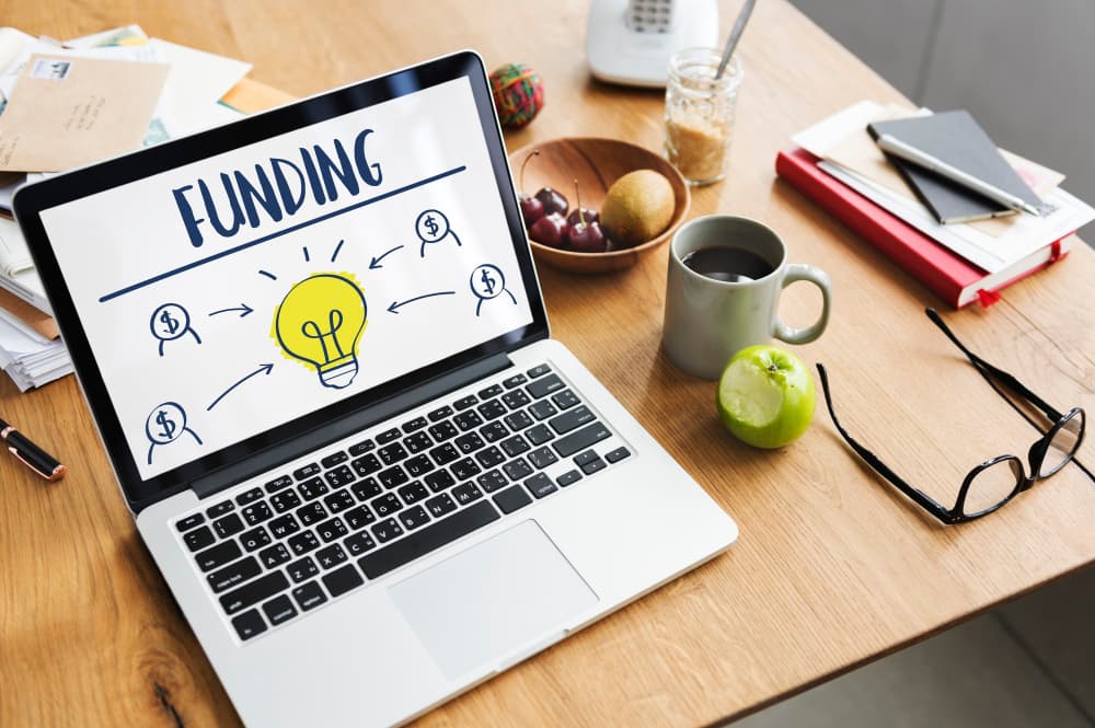 Ein Laptop zeigt eine Darstellung zur Finanzierung, auf der eine leuchtende Glühbirne und Strichmännchen mit Dollarzeichen in ihren Köpfen abgebildet sind.