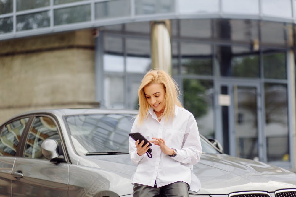 Eine Frau lehnt sich gegen die Motorhaube eines Autos und tippt auf ihrem Smartphone.