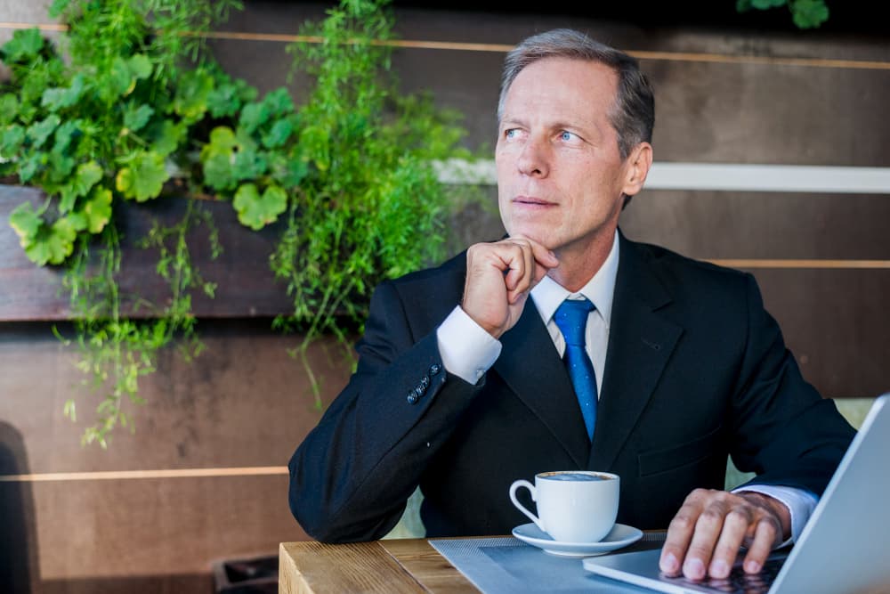 Ein Mann in Anzug sitzt nachdenklich an einem Tisch mit einer Tasse und einem Laptop.