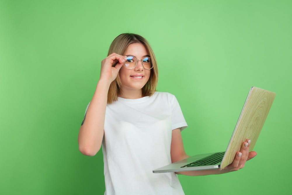 Eine Frau mit Brille und Laptop steht vor einer grünen Wand.
